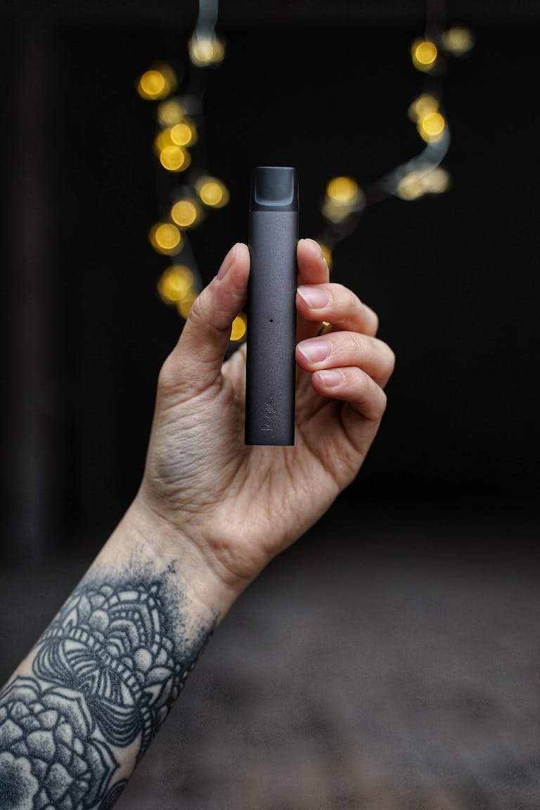 A tattooed hand holds a sleek vape pen against a dark bokeh background.