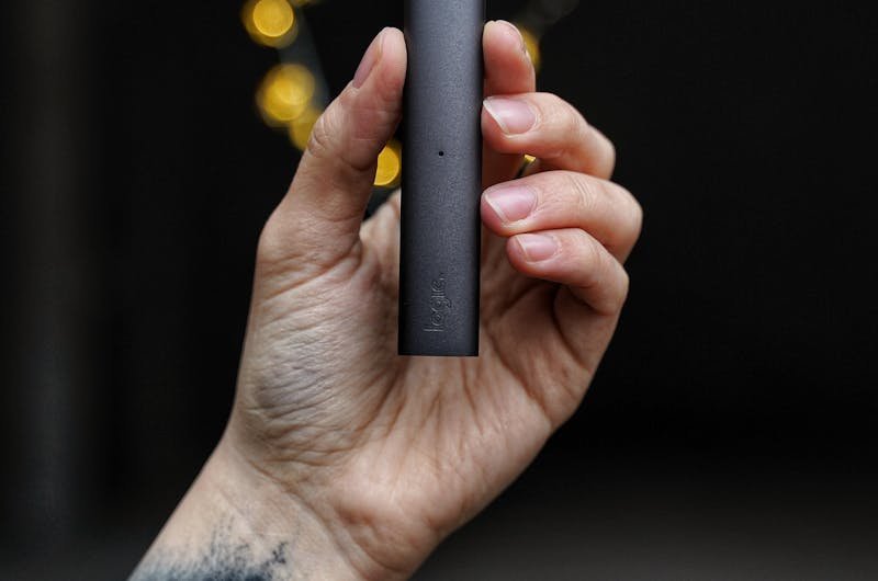 A tattooed hand holds a sleek vape pen against a dark bokeh background.