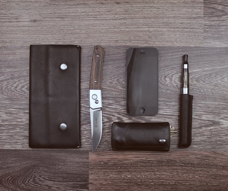 Flat lay of modern lifestyle items including a knife, smartphone, and vape on a wooden surface.