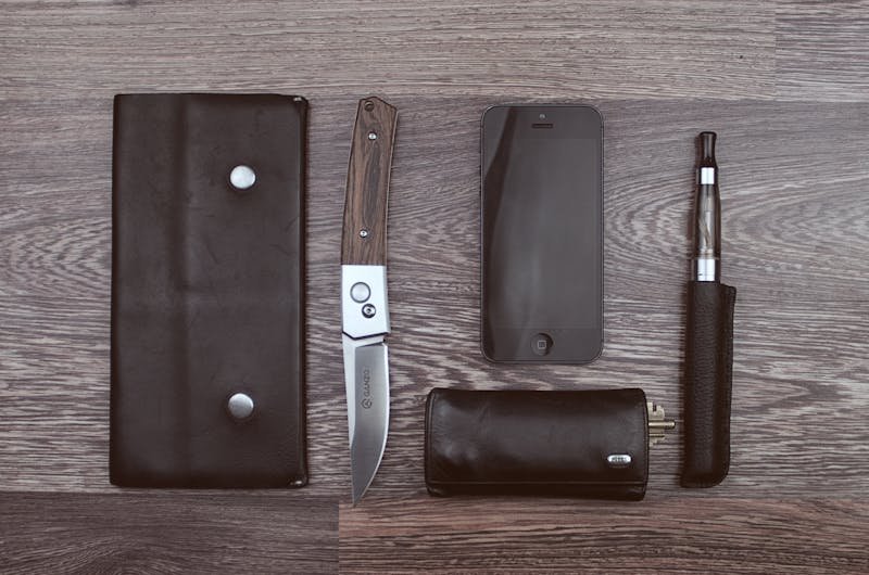 Flat lay of modern lifestyle items including a knife, smartphone, and vape on a wooden surface.