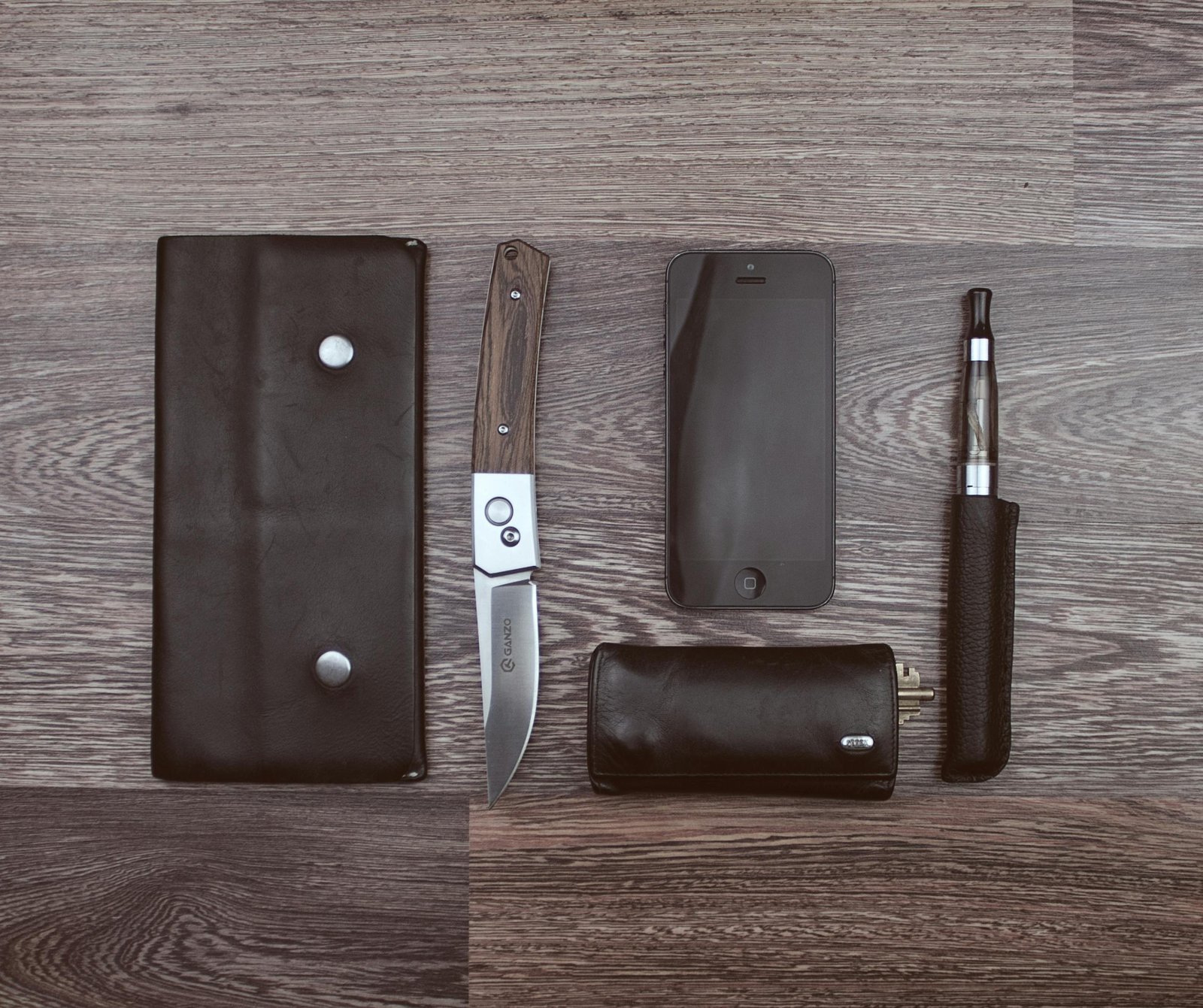 Flat lay of modern lifestyle items including a knife, smartphone, and vape on a wooden surface.